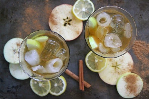 2 glasses filled with ice and a cocktail with apple slices and cinnamon sticks scattered around it. 