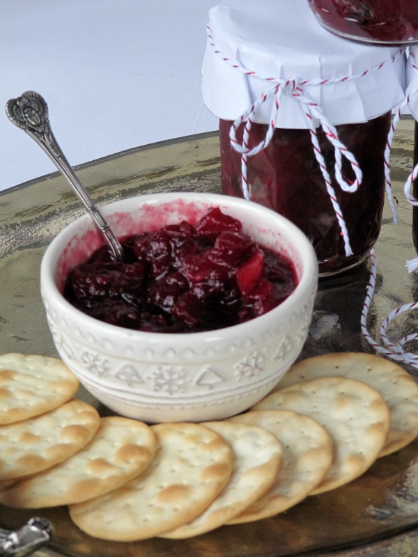 Cranberry apple chutney in a white bowl and a jar with water crackers scattered around it. 