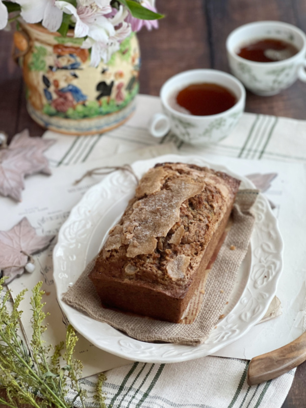 pumpkin Zucchini nut bread on a white oval plate with 2 cups of tea and a colorful bucker of flowers.