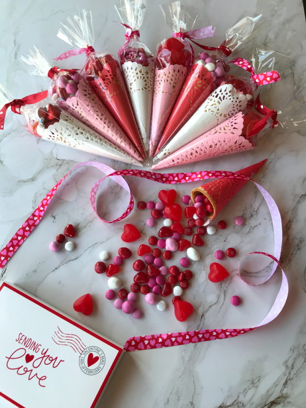 Red, white and pink paper cones filled with valentines day candy in an arc with a ribbon wrapped at the bottom and spilled candy.