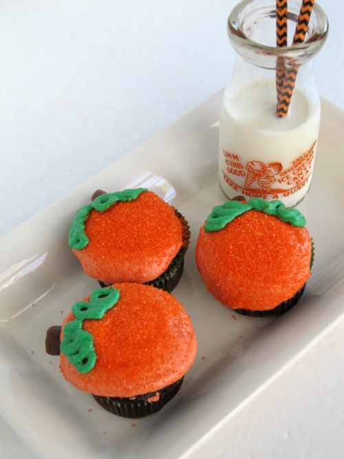 Three cupcakes decorates with orange and green frosting to look like pumpkins on a white platter with a small pink of milk