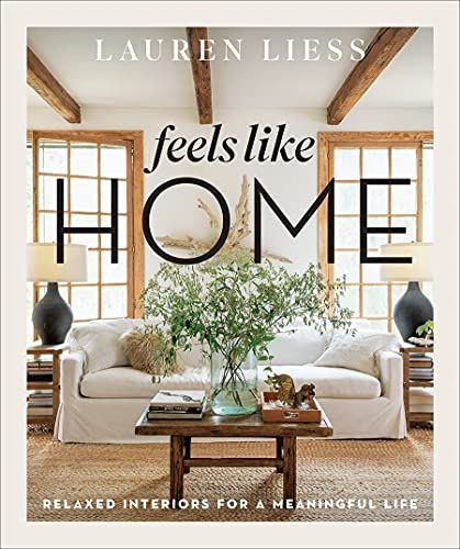 book cover of Feels like home the view of a white couch with in front of two windows with a table in front that has a large plant on it and two lamps on ash side