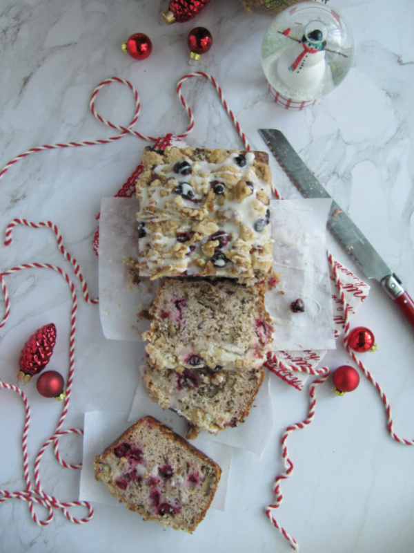 cranberry walnut loaf wrapped in white paper and tied with red and white string on a white piece of marble with scattered christmas bulbs a snow globe and string