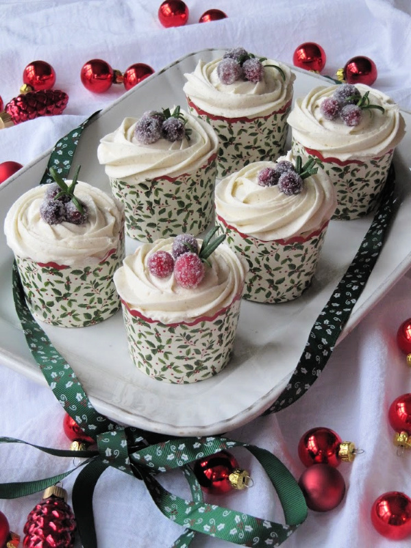 Sic cupcakes in green and red paper cups with white icing and candies cranberries on top sitting on a white tray with red and green ornaments scattered around