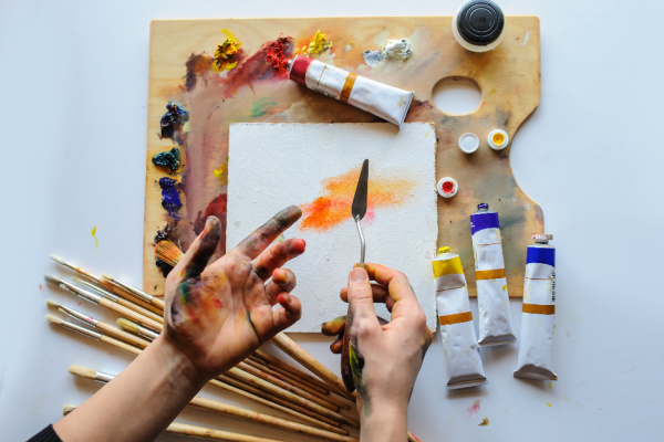 artist pallet and a variety of tubes of paint along with brushes and a pair of hands holding a pallet knife 