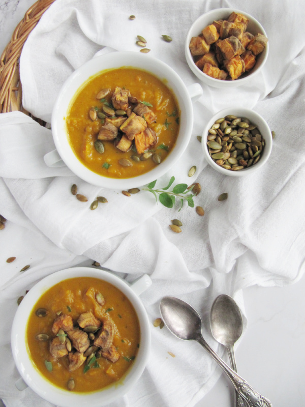 Sweet Potato and Pumpkin Soup in two white bowls with handles and a small bowl of pumpkin seeds and roasted sweet potatoes on the side.