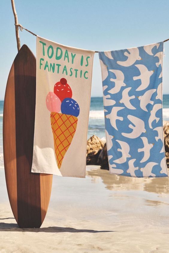 Beach towels, one with an ice cream cone and the other blue with white doves hanging on a line next to a brown surf board. 