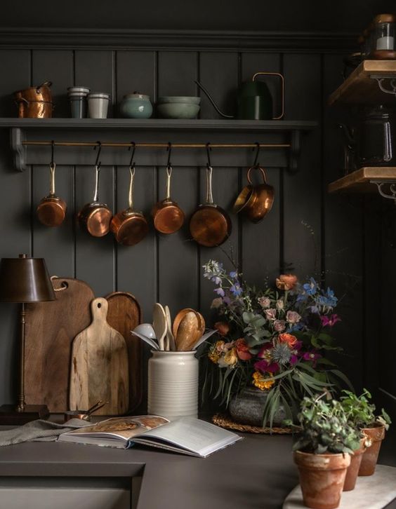 Dark green bead board wall with hanging copper pots above a counter with wood cutting boards and flowers.