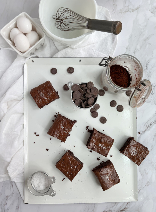 sweetened condensed milk brownies on a white pan with a basket of eggs a white bowl and whisk and a jar of cocoa powder.