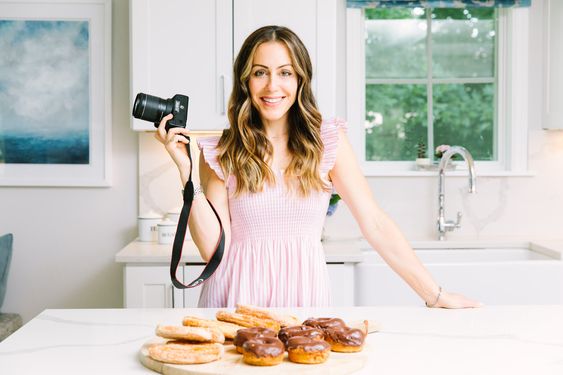 Kat Ashmore in a pink dress holding a camera in her all white kitchen standing in front of a plate of donuts. 