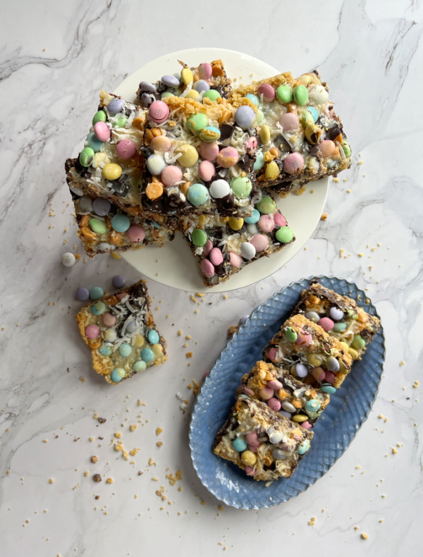 Easter M&M 7 Layer Bars on a blue oval tray and a white cake platter and one with a bite out sitting on the side.